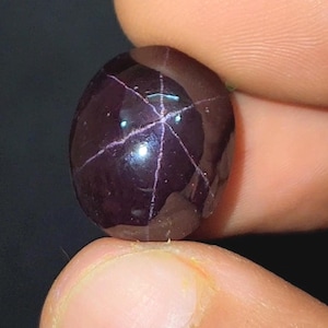May include: A polished, oval gemstone in a deep burgundy hue. It displays a six-rayed star pattern across its surface, with pale lines radiating from the centre. The gemstone is held between two fingers against a dark backdrop.