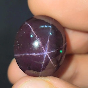 May include: A deep purple, oval gemstone with a polished surface. The stone displays a star-shaped pattern of lighter lines. The gem is held between fingers, highlighting its smooth, reflective appearance. A striking piece of jewellery.