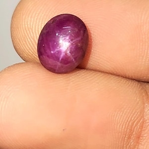 May include: A polished, oval-cut, deep red gemstone exhibiting a star effect. The surface reflects light, forming a six-pointed star. The gem is held between fingers, highlighting its colour and shape.