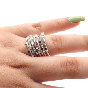 Peut inclure: Une bague en argent composée de plusieurs anneaux, chacun orné d'une série de pierres précieuses colorées. Les pierres précieuses comprennent des pierres rouges, bleues, vertes et transparentes. La bague est portée sur un doigt.