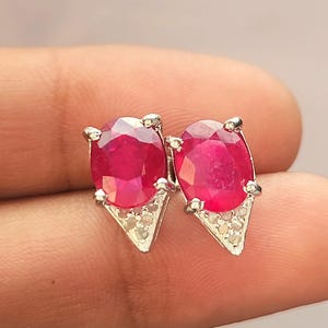 Può includere: Un paio di orecchini di rubino. Ogni orecchino presenta un grande rubino rosso scuro a taglio ovale incastonato in un metallo color argento. I rubini sono montati su una base triangolare con piccole pietre chiare.