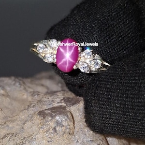 Peut inclure: Bague en or ornée d'un grand rubis étoilé ovale. Le rubis est d'un rose profond et présente un motif en étoile à six rayons. La bague est ornée de petits diamants clairs de chaque côté du rubis.