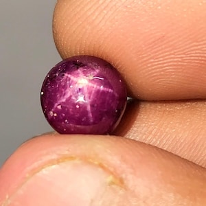 May include: A spherical, polished, deep magenta gemstone, held between fingers. The gem shows a six-rayed star pattern across its surface, with a glossy finish. The gem is a star ruby, a type of corundum.
