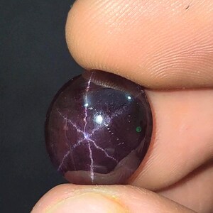 May include: A polished, oval-shaped, dark purple gemstone. The gem has a star-like pattern of light-coloured lines radiating from the centre. The stone is held between two fingers against a dark background.