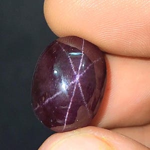 May include: A polished, oval-shaped, dark purple gemstone. The gem has a six-rayed star pattern across its surface. The stone is held between two fingers, with a dark background.