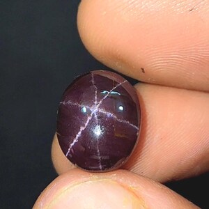May include: A polished, oval gemstone in a deep burgundy hue. The stone displays a star-like pattern of white lines across its surface. The gem is held between fingers against a dark backdrop.