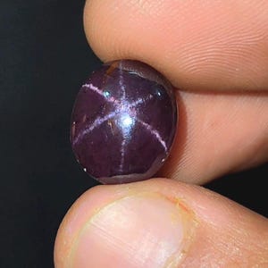 May include: A deep purple, oval gemstone with a star pattern. The six-rayed star is visible on the surface, with white lines radiating from the centre. The gem is held between two fingers against a dark background.