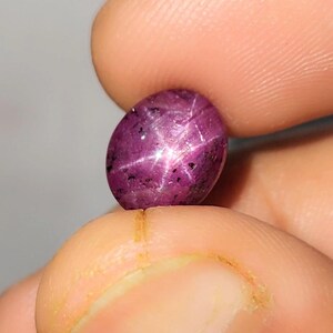 Può includere: Pietra preziosa di rubino ovale e lucidata con un effetto stella a sei raggi. La gemma è di un colore rosso-violaceo intenso, con una superficie liscia e riflettente. L'effetto stella è visibile sulla superficie.