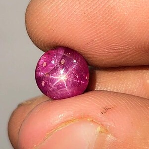May include: A polished, round ruby gemstone exhibiting a star effect. The gem is a deep pinkish-red hue, displaying a six-rayed star pattern across its surface. The gemstone is held between two fingers.