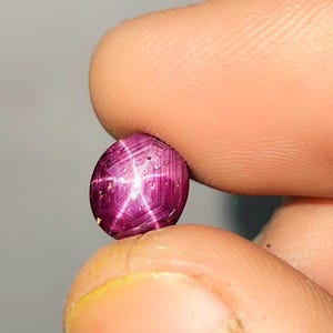 Può includere: Una gemma di rubino lucidata, di forma ovale, con un effetto stella a sei raggi. La gemma è di un colore rosso intenso, con un motivo a forma di stella che si irradia dal centro. La gemma è tenuta tra due dita.