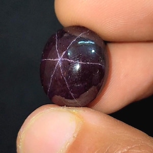 May include: A dark purple gemstone with a star-like pattern of light lines. The oval-shaped gem is polished, reflecting light. It is held between fingers, highlighting its colour and design. The gem's surface has a smooth, glossy appearance.