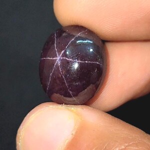 May include: A dark, oval gemstone with a star-like pattern of light lines. The gem is held between fingers, showing its polished, reflective surface and deep burgundy colour. This gemstone is a unique piece of jewellery.