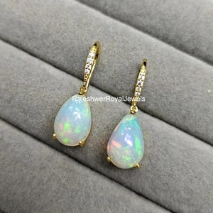 Könnte beinhalten: Ein Paar goldfarbene Ohrhänger mit tropfenförmigen Opalsteinen. Die Ohrringe haben eine kleine Reihe klarer Steine am oberen Teil. Die Opale weisen eine Mischung aus Farben auf, darunter Grün, Rosa und Blau.
