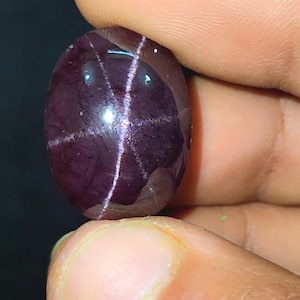 May include: A deep burgundy, oval gemstone with a star-shaped pattern of white lines. The polished stone is held between fingers, highlighting its reflective surface. The gem's colour is a rich, dark red, and the stone is smooth to the touch.