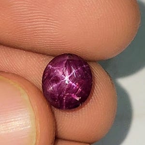 Könnte beinhalten: Ein runder, tiefroter Edelstein mit einem sternförmigen Muster auf der Oberfläche. Der Stein wird zwischen Fingern gehalten, wodurch seine polierten, reflektierenden Facetten hervorgehoben werden. Der Sterneffekt wird durch Einschlüsse im Stein erzeugt.