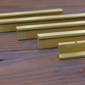 Puede incluir: Varias barras metálicas doradas con perfil rectangular dispuestas sobre una superficie de madera oscura. Las barras tienen una ranura empotrada y son de diferentes longitudes. Se aprecia la veta de la madera.