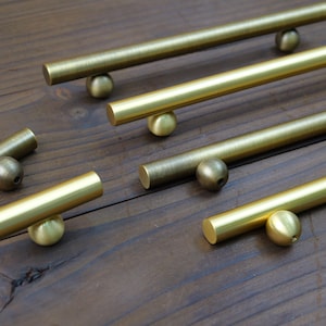 May include: Several brass cabinet pulls and knobs are arranged on a wooden surface. The pulls are cylindrical with a spherical base, and the knobs are small spheres. The hardware has a warm, golden hue.