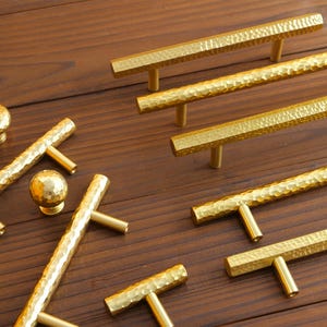 May include: A selection of gold-coloured cabinet hardware, including knobs and bar pulls. The hardware has a hammered texture and is arranged on a dark wood surface. The knobs are round and the pulls are rectangular with T-shaped posts.