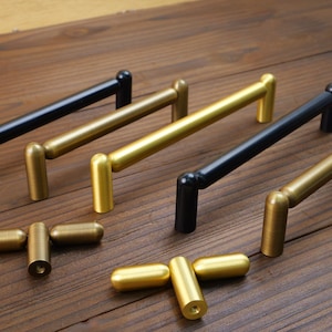 May include: A collection of cabinet pulls in gold and black. The pulls have a cylindrical bar design with rounded ends and are arranged on a wooden surface. Some pulls are gold, while others are black.