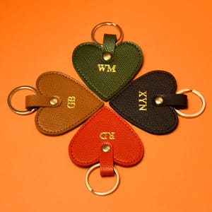Könnte beinhalten: Vier herzförmige Schlüsselanhänger in symmetrischer Anordnung. Jeder Schlüsselanhänger hat eine andere Farbe: Braun, Grün, Schwarz und Rot. Jeder Schlüsselanhänger hat einen goldfarbenen Schlüsselring und goldfarbene Initialen. Der Hintergrund ist orange.