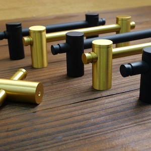 May include: A collection of modern cabinet pulls in gold and black. The pulls have a cylindrical design with a T-shaped handle. The hardware is made of metal and is displayed on a wooden surface. The pulls are designed for use on drawers and cabinets.