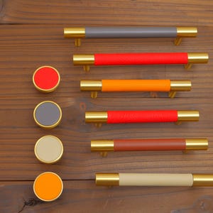 Puede incluir: Un conjunto de herrajes para armarios, que incluye tiradores y pomos redondos. Los tiradores tienen extremos dorados y centros envueltos en cuero de colores (gris, rojo, naranja y marr&oacute;n). Los pomos tienen colores a juego.