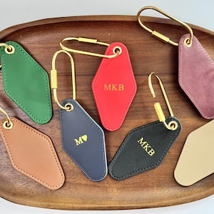 May include: A selection of personalised keyrings in shades like green, red, and black, displayed on a wooden tray. Each keyring has a diamond shape, gold-coloured hardware, and initials.