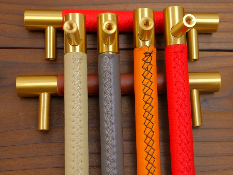 Puede incluir: Cuatro tiradores de armario de lat&oacute;n con asas de cuero de colores. Los mangos son beige, gris, naranja y rojo, con costuras decorativas. Los tiradores tienen un dise&ntilde;o en T y est&aacute;n colocados sobre un fondo de madera oscura.