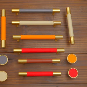 Puede incluir: Una colecci&oacute;n de herrajes para armarios, que incluye tiradores y pomos para cajones. Los tiradores presentan extremos met&aacute;licos dorados y secciones centrales envueltas en cuero de colores en tonos grises, beige, naranja, rojo y marr&oacute;n. Tambi&eacute;n se incluyen pomos redondos.