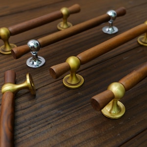 May include: Several wooden cabinet pulls with brass and silver-toned hardware are arranged on a dark wood surface. The pulls have a cylindrical wooden handle and a decorative base. The brass hardware has a gold finish.