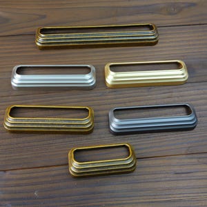 May include: A collection of rectangular metal drawer pulls in various finishes, including antique brass, silver, and gold. Each pull has a recessed center and a layered, decorative design. The pulls are arranged on a wooden surface.