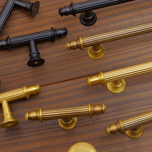 Puede incluir: Una variedad de herrajes para gabinetes, que incluyen manijas y perillas en tonos negros y dorados. Las manijas presentan una variedad de diseños, incluyendo tiradores de barra y en forma de T, y están dispuestas sobre una superficie de madera. El hardware es probablemente para gabinetes de cocina o baño.