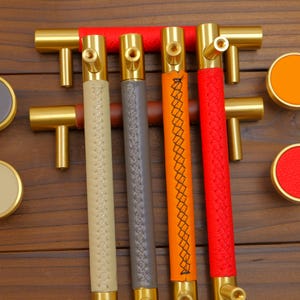 Puede incluir: Un conjunto de herrajes para armarios, que incluye tiradores en T de lat&oacute;n y pomos redondos. Los tiradores tienen empu&ntilde;aduras forradas de cuero en beige, gris, naranja y rojo. Los pomos son grises, beiges, naranjas y rojos con bordes de lat&oacute;n.