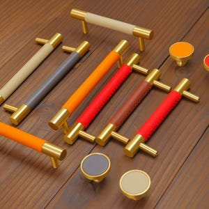 Puede incluir: Un conjunto de tiradores y pomos para armarios. Los tiradores tienen extremos dorados y empu&ntilde;aduras de cuero en beige, gris, naranja, rojo y marr&oacute;n. Los pomos tienen tapas de colores a juego. Medidas en cent&iacute;metros.