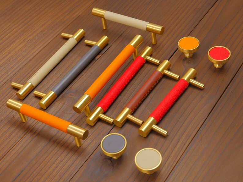 Puede incluir: Un conjunto de tiradores y pomos para armarios. Los tiradores tienen extremos dorados y empu&ntilde;aduras de cuero en beige, gris, naranja, rojo y marr&oacute;n. Los pomos tienen tapas de colores a juego. Medidas en cent&iacute;metros.