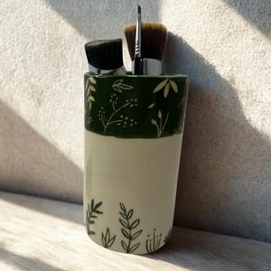 May include: A ceramic makeup brush holder with a green floral design. The holder is cylindrical, with a cream-coloured base and a dark green top section. Three makeup brushes are visible inside the holder.