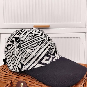 May include: A black and white patterned baseball cap with a black brim. The cap features a geometric design and is displayed on a woven basket.