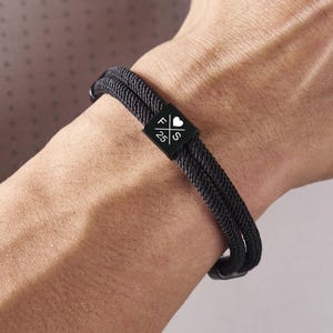 Op de afbeelding: Zwart touw armband met een vierkant, zwart metalen accent. Het accent heeft een wit hart symbool en de letters "FS" en "25". De armband wordt om een pols gedragen.
