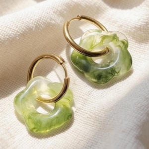 Puede incluir: Un par de pendientes de aro dorados con colgantes florales translúcidos verdes y blancos. Los pendientes se exhiben sobre una tela blanca texturizada, con una iluminación suave.