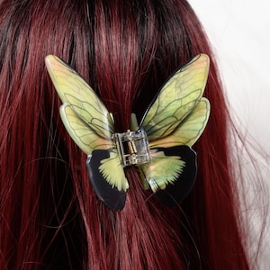 May include: A butterfly hair clip with translucent green and black wings, attached to a clear plastic clip. The clip is set against a backdrop of deep red hair.