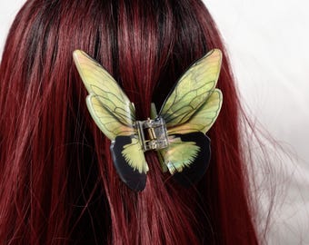 May include: A butterfly hair clip with translucent green and black wings, attached to a clear plastic clip. The clip is set against a backdrop of deep red hair.