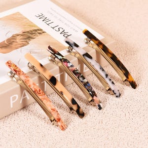 Puede incluir: Seis pinzas para el pelo decorativas con varios diseños y colores, incluyendo carey, mármol y floral. Las pinzas están colocadas sobre un libro con la palabra "PAST TIME" visible.