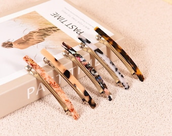May include: Six decorative hair clips in various patterns and colors, including tortoiseshell, marble, and floral designs. The clips are arranged on a book with the word "PAST TIME" visible.