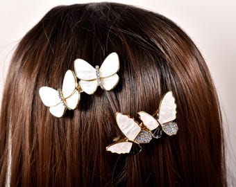 Puede incluir: Tres pinzas para el pelo con forma de mariposa. Las pinzas presentan alas blancas con marcos dorados y están adornadas con pequeños cristales brillantes. Los accesorios para el cabello se colocan sobre un fondo de cabello castaño.