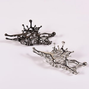 Puede incluir: Dos pinzas de pelo abstractas de metal. Una es negra, la otra plateada. Cada pinza tiene un diseño orgánico único con forma de salpicadura. Las pinzas se muestran sobre un fondo blanco.