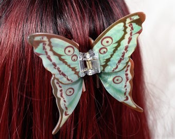 May include: A butterfly-shaped hair clip with a clear plastic clasp. The butterfly wings are light green with brown and white details. The clip is attached to red hair.