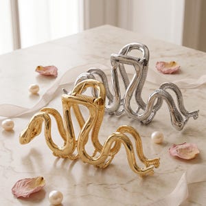 May include: Two metal hair claw clips, one in a polished gold tone and one in a polished silver tone. Each clip features a detailed, winding snake design that forms the structure of the claw, with a spring mechanism at the top for opening and closing.