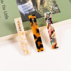 Può includere: Tre fermagli per capelli rettangolari. Il primo è bianco sporco con testo dorato "CLARA". Il secondo è ambrato con macchie nere e il testo "Evelyn". Il terzo è multicolore con rosa, verde e nero.