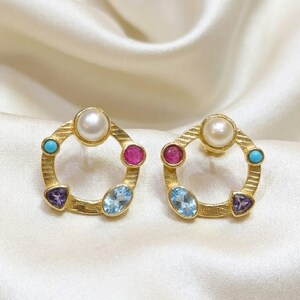 May include: A pair of gold-toned earrings, each featuring a central pearl, surrounded by a turquoise, ruby, blue topaz, and amethyst gemstone. The earrings have a circular design with textured detailing.