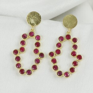 May include: A pair of gold-coloured earrings. Each earring has a circular top and a teardrop-shaped design, adorned with small, round, red gemstones. The earrings are presented on a white surface.